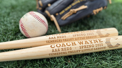 Two wooden baseball bats with engraved text on a grass background, accompanied by a baseball and a black baseball glove.